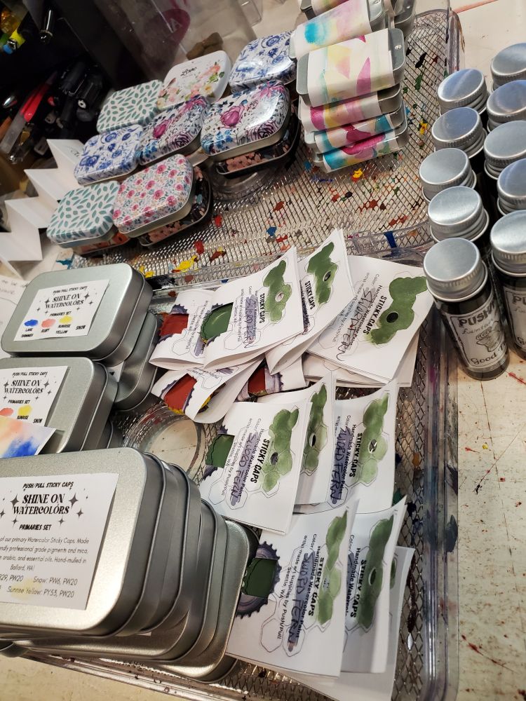 Stack of store-made art supplies (mostly watercolors in both tins and individual caps, a few bottles of ink) on the front desk.