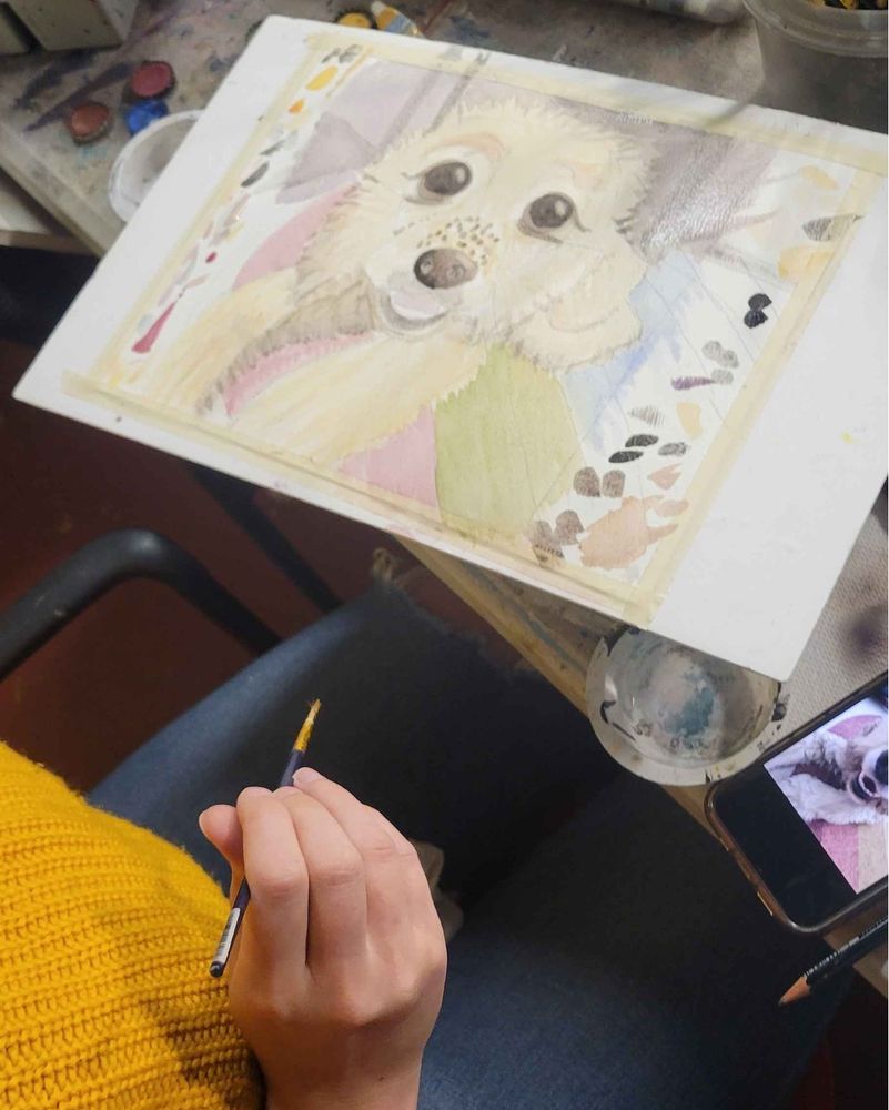 Photo of an in-progress watercolor painting of a small dog. Artist hand holding paint brush in foreground.