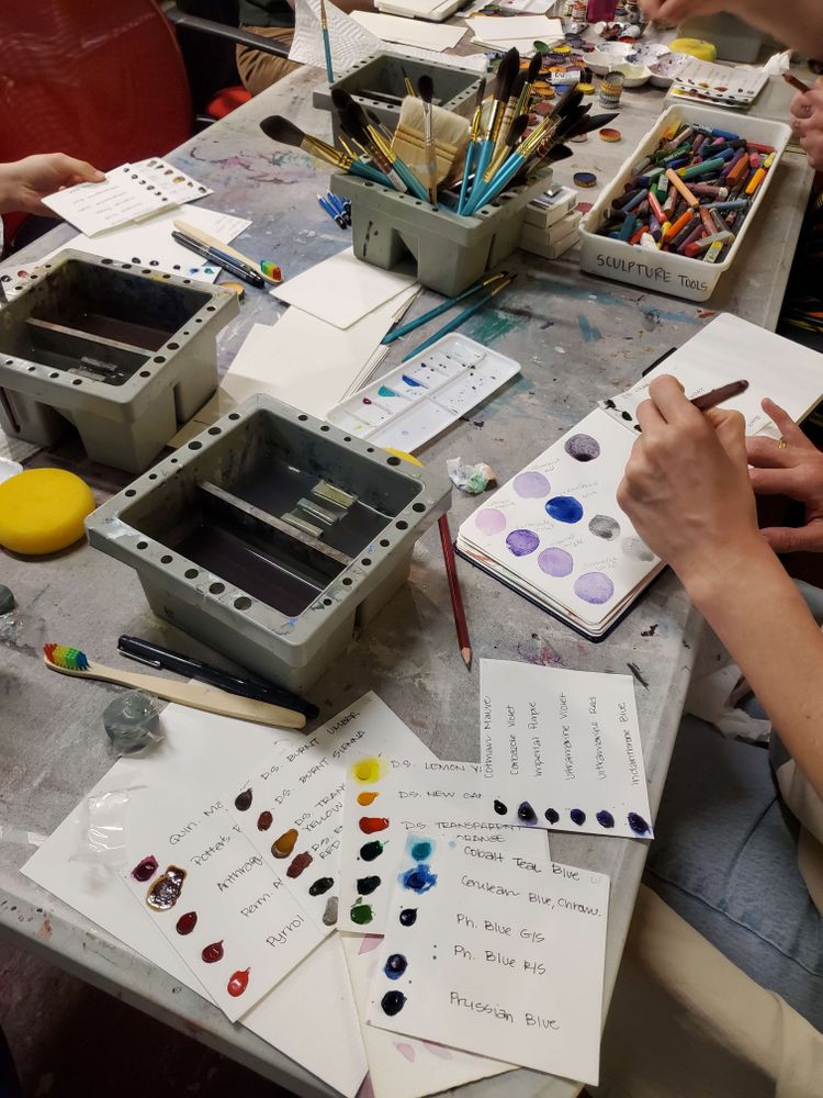 Messy art table covered in watercolor supplies; at the front are various dots of color in samples/swatches. Two people are working on various swatches in mini sketchbooks.