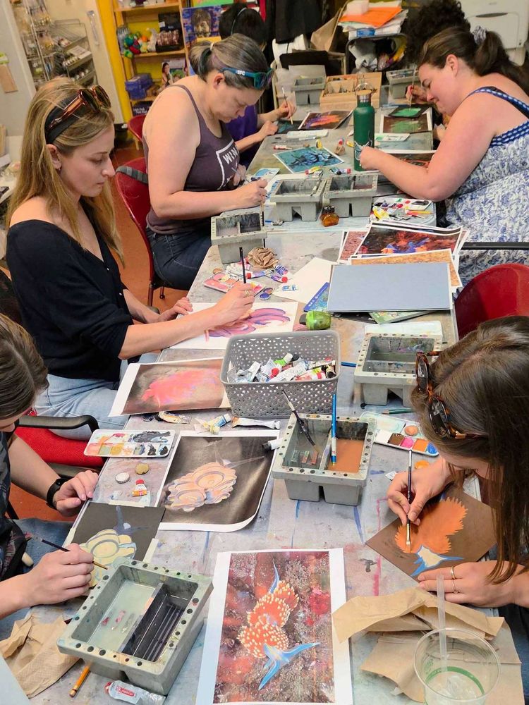 Several people at a messy art table with lots of printed reference of nudibranches, working on gouache paintings.