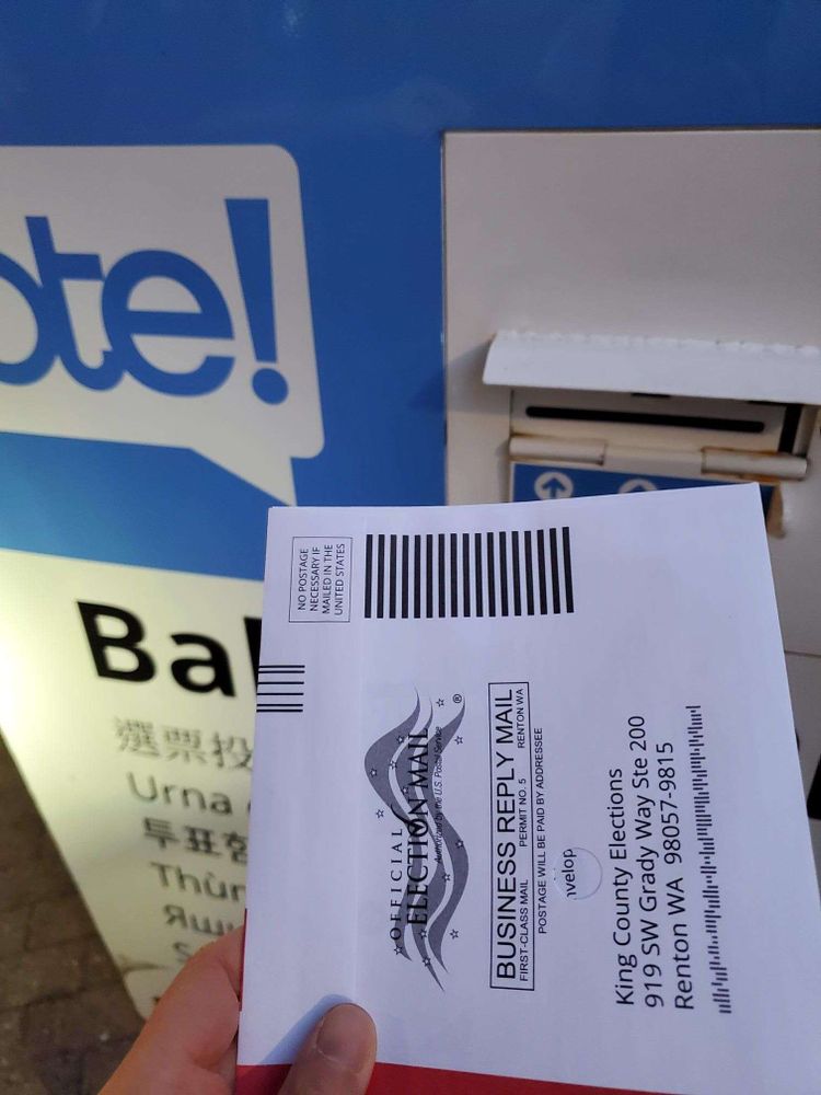 Hand holding a ballot envelope, about to insert it into a ballot drop box.