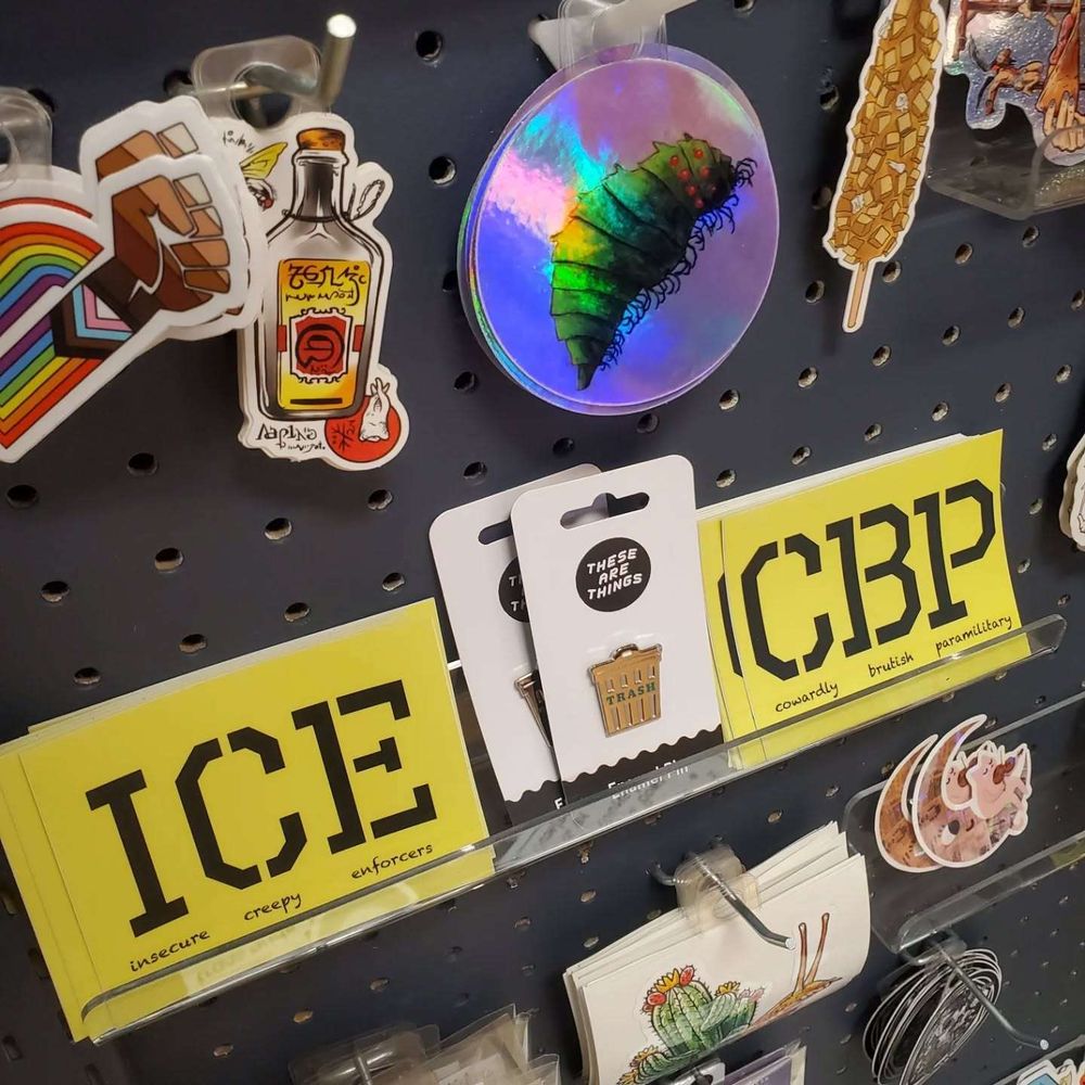 Close up of sticker corner in store, focus on two yellow stickers with a trash can enamel pin between them. One sticker reads "ICE, insecure creepy enforcers" and the other reads "CBP, cowardly brutish paramilitary."