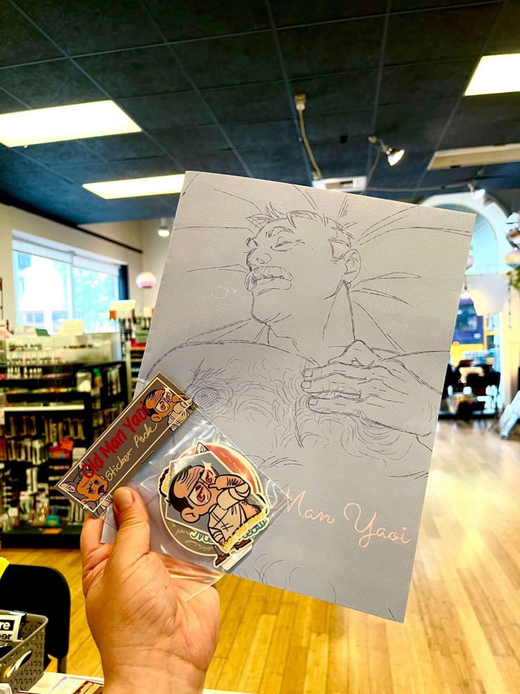 Hand olding up a book featuring an older shirtless man sleeping and a small sticker pack featuring chibis. The sticker pack reads "Old Man Yaoi." The book says the same, though the "Old" is obscured by the sticker pack.