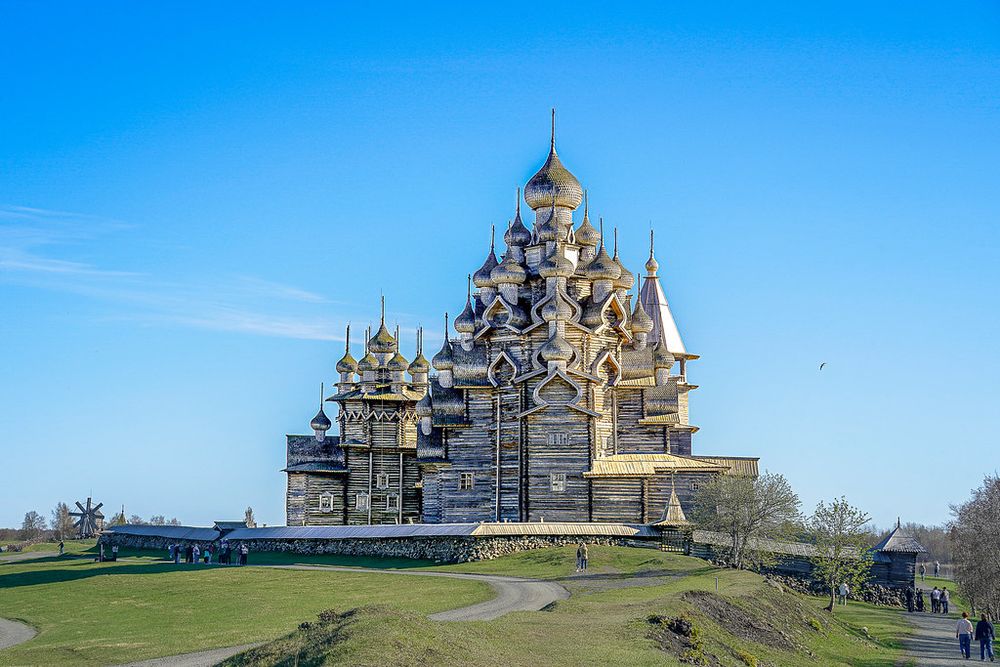 Ensemble of the stunning unique wooden churches, built without nails, with intricate onion domes stands majestically against a clear blue sky on a grassy landscape. People can be seen walking around the historic site, suggesting its cultural significance and tourist appeal. This is the Kizhi Pogost a unique monument of Russian wooden architecture (15th-18th centuries), a universally recognized masterpiece of world architecture.


