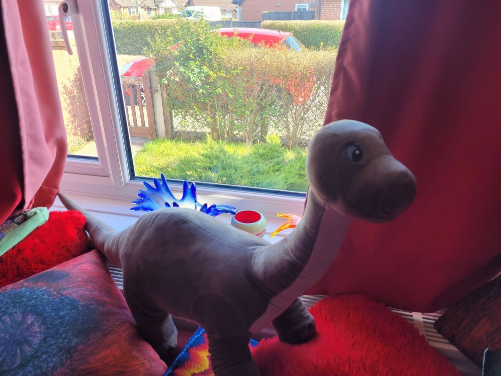 A 90cm long IKEA Jattelik dinosaur/brontosaurus standing on pillows in front of a window. It is light brown with cream undersides.