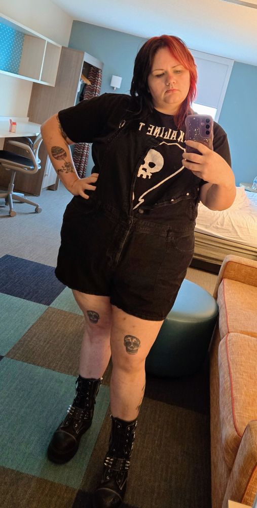 A femme person stands with hand on her hip, wearing an Alkaline Trio t shirt and shortalls. She's wearing platform boots and a sassy expression