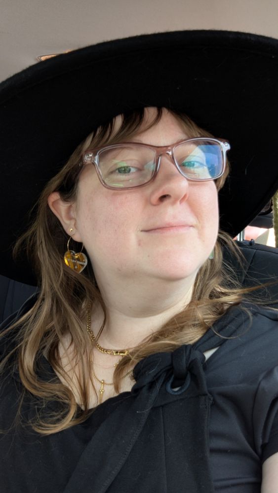 A blonde white woman in a big black sunhat and black dress with heart earrings 