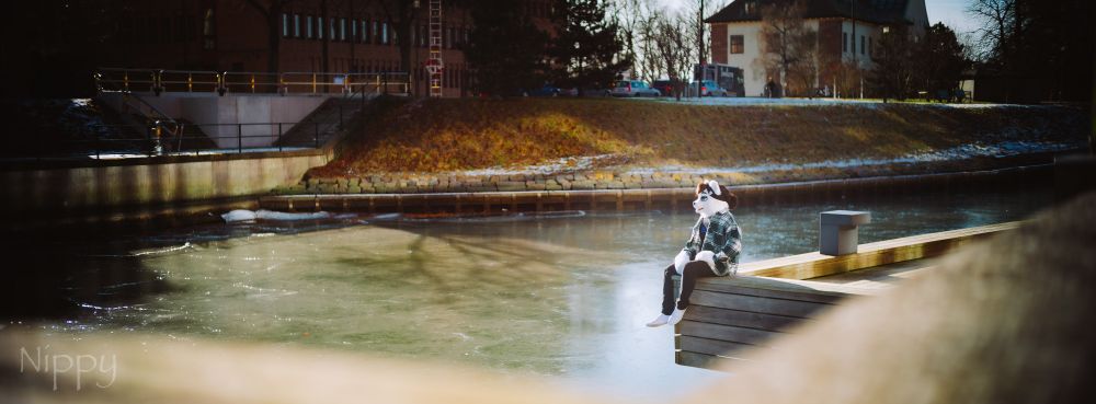 A wide angle scenic photo with an ermine partial fursuiter. The fursuiter is sitting on a pier's edge just slightly right of the photo's centerline. He's wearing a black beanie, green and white plaid shirt, black jeans and white shoes. He's looking away from the camera, towards the left side of the frame. A narrow channel of water and its opposing bank is seen in the background, along with some red and white brick buildings.