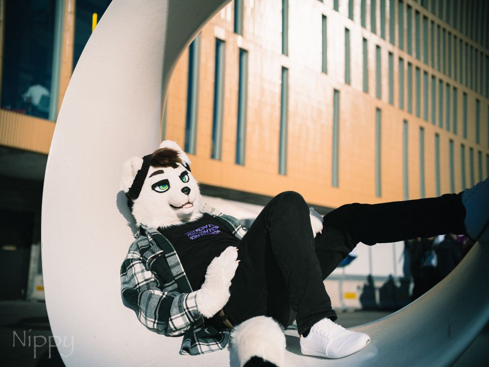 An ermine partial fursuiter laying on a white sculpture. The fursuiter is wearing a black beanie, dark green plaid shirt over a black t-shirt, black jeans and white shoes. A light brown wall with long vertical windows can be seen in the background.