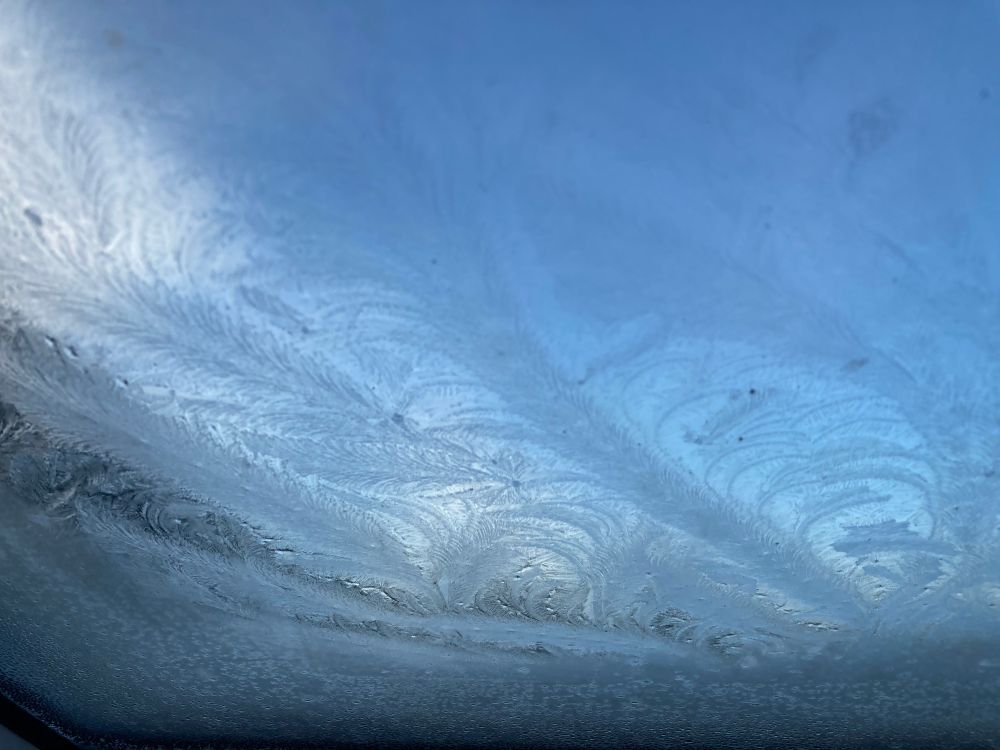Swirly feathered ice crystal patterns frozen on a window with blue sky peeking through behind 