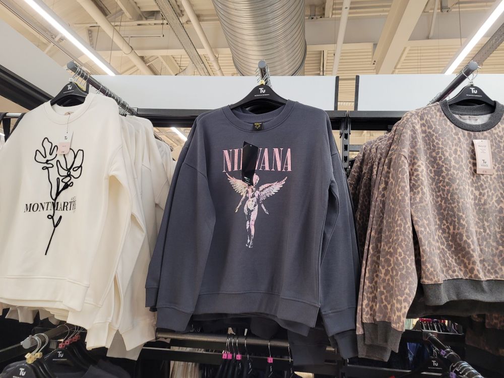A range of sweatshirts for middle aged women hang on a rack in Sainsburys. The middle one is Nirvana merch.