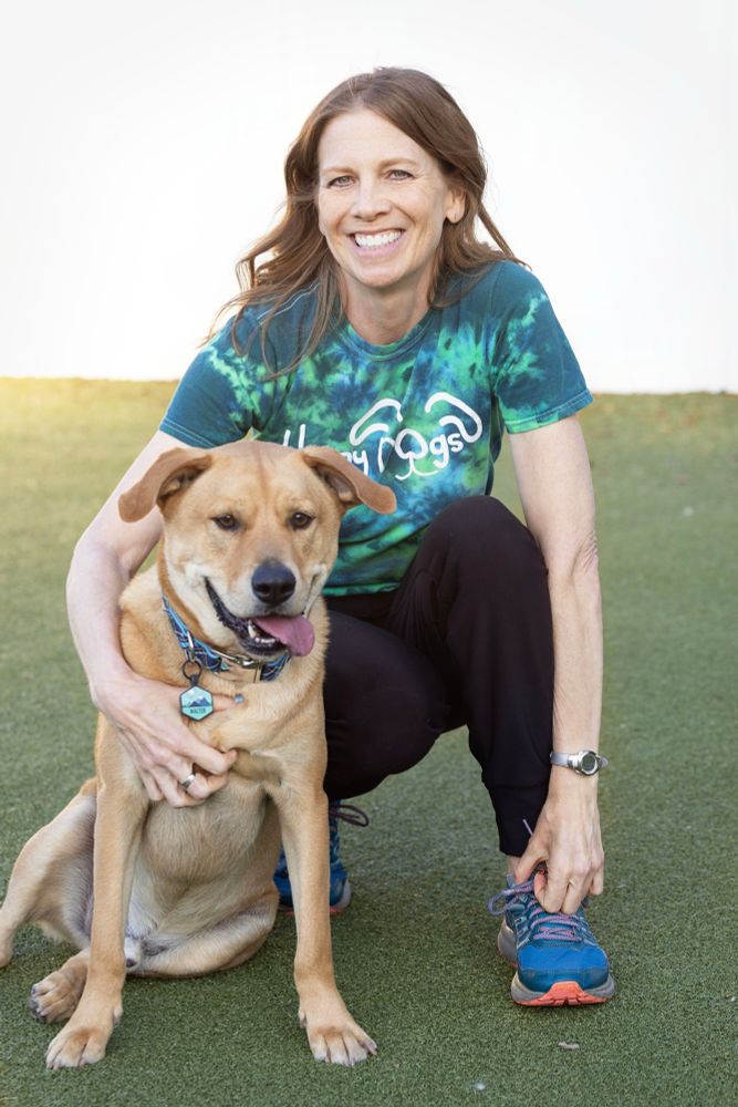 A white, red-haired woman is smiling and squatting next to a light brown lab-mix dog. 