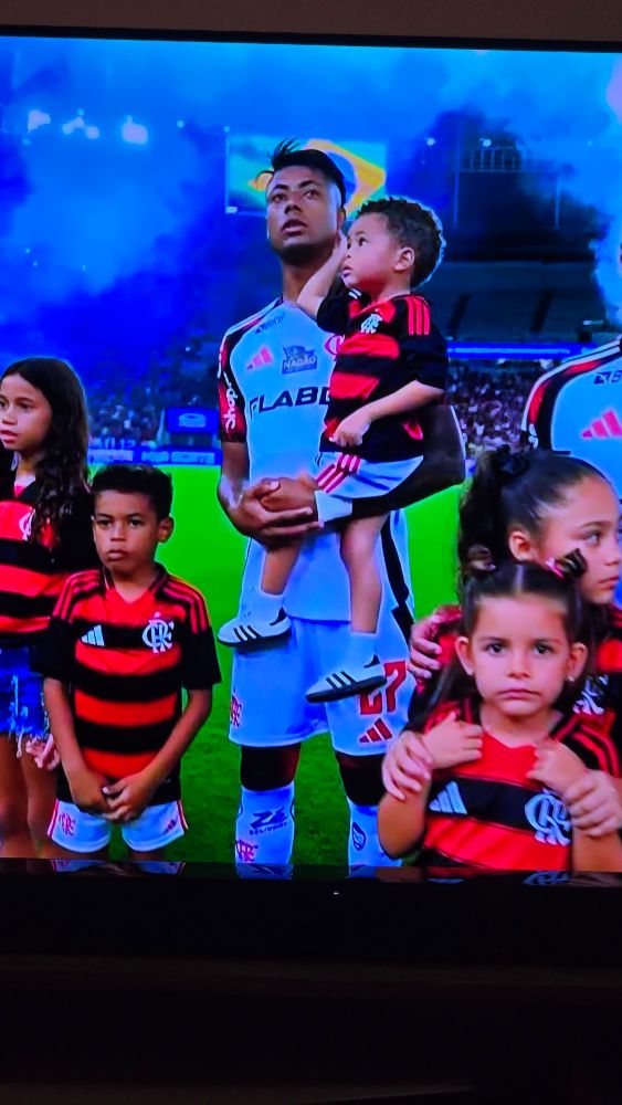 Fotografia de uma televisão mostrando jogadores de futebol do time do Flamengo cercados de criancas. Em destaque o atleta Bruno Henrique com um bebê no colo