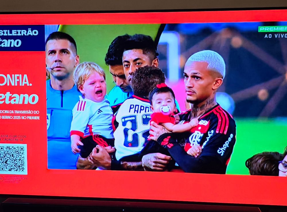 Jogadores do time de futebol Flamengo segurando crianças no colo. Em destaque os atletas Bruno Henrique e Wesley segurando bebês.