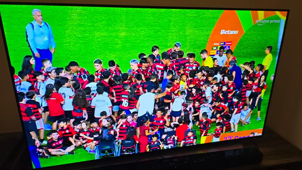 Fotografia de uma televisão mostrando o início de um jogo de futebol, com.dezenas de crianças aglomeradas ao redor dos jogadores do time do Flamengo.