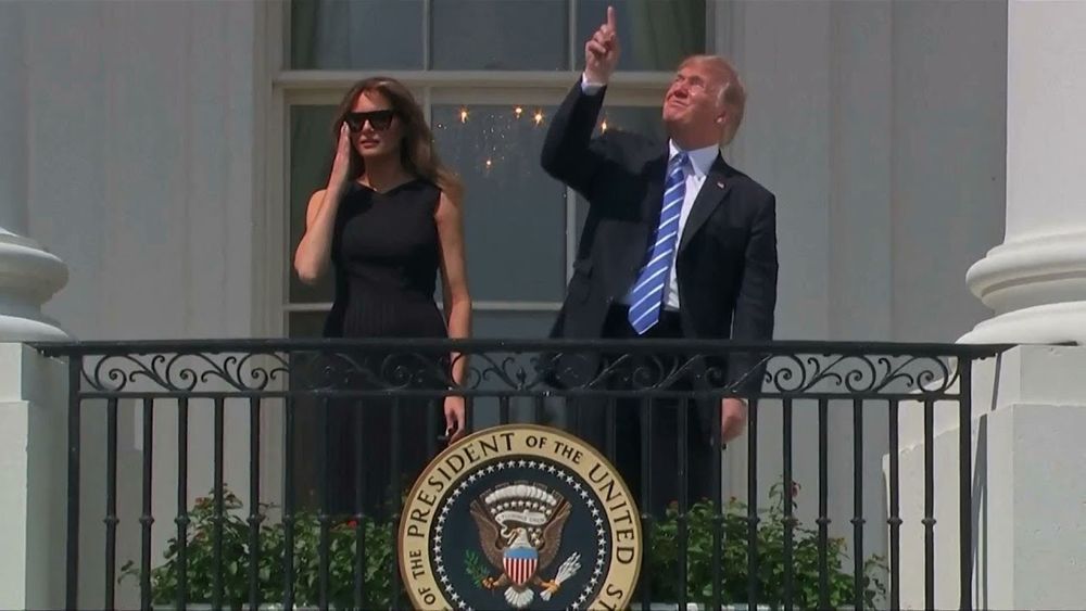 Trump staring and pointing at the solar eclipse without protective glasses, and Melania wearing the glasses and looking ahead.