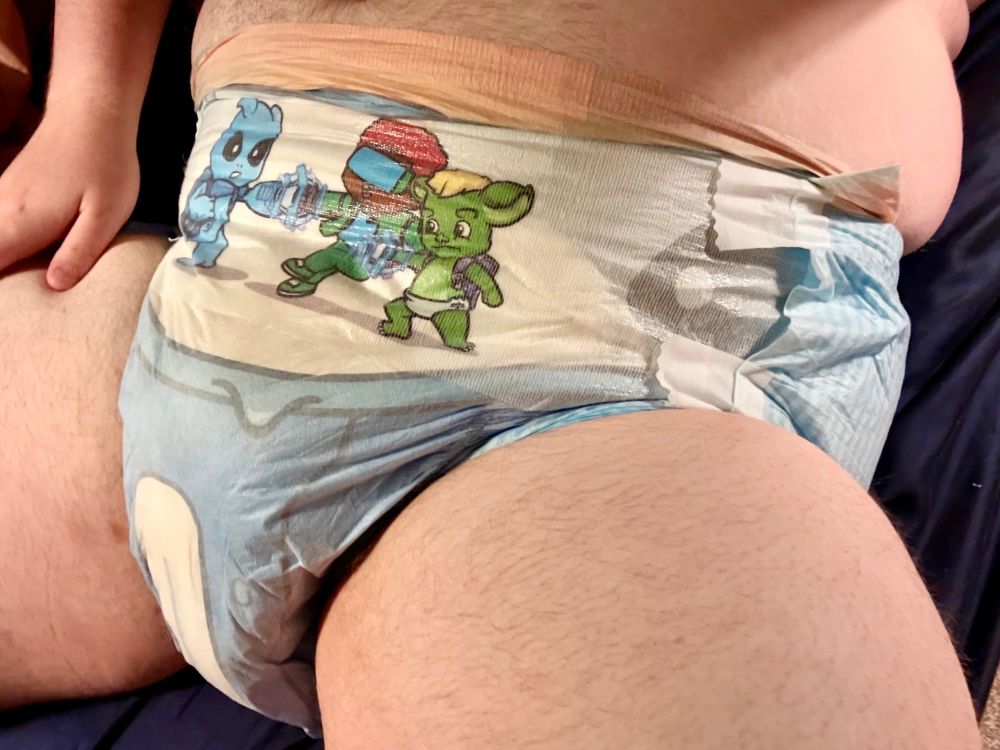 A chubby man lying down wearing a very wet blue adult diaper with cartoon aliens on it