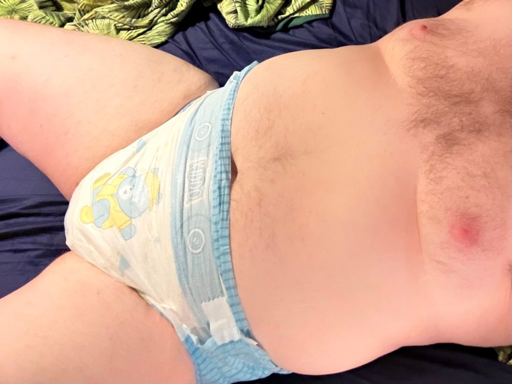 A chubby hairy man lying in bed wearing just a wet adult diaper. The diaper is light blue and white with a pattern showing buttons and a cartoon bear in a yellow overcoat jumping in a puddle