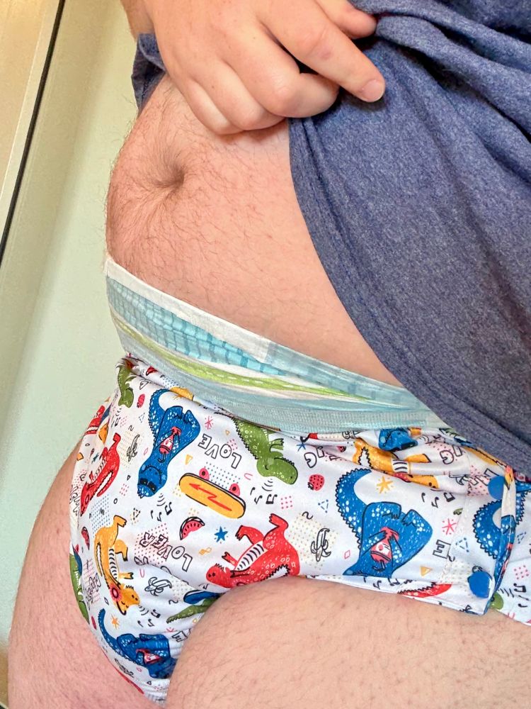 A chubby man pulling up his T-shirt to show a dino-print diaper cover over a very wet adult diaper