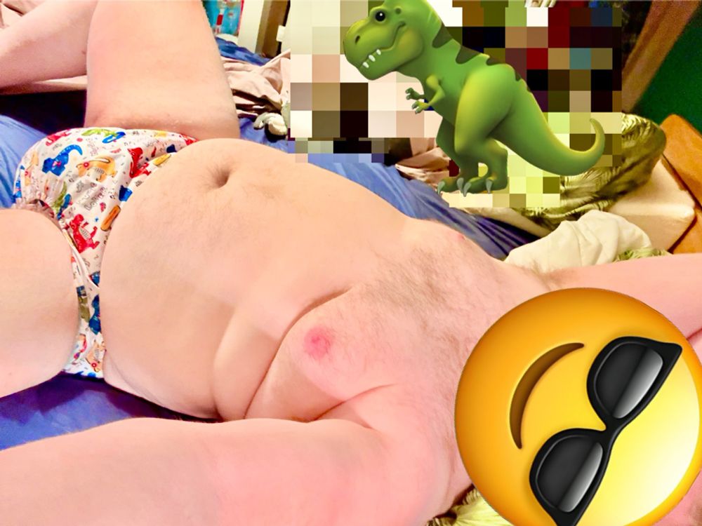 A chubby man lying in bed naked except from a dino-print diaper cover with a wet looking diaper underneath.