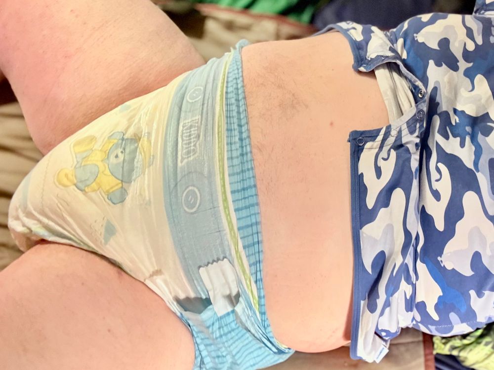 A chubby man lying on a changing mat with a blue camo romper suit pulled up showing a very wet adult diaper with a cartoon pattern