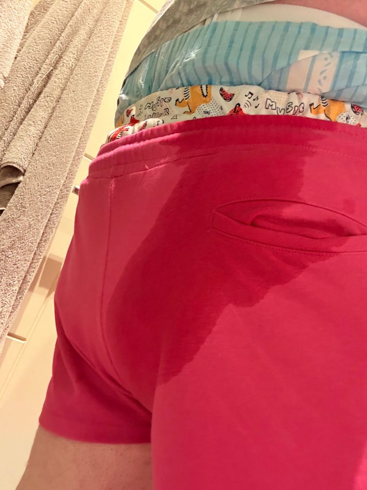 A chubby man standing in a pair of bright pink shorts with a large wet patch down the back. A dino-print diaper cover and a baby blue adult diaper is peeking over the top of his shorts.