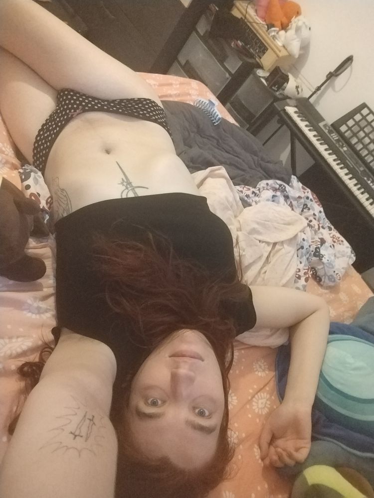 Selfie d'une meuf aux cheveux rouges allongée en culotte sur son lit avec un crop top noir