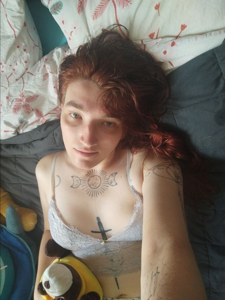 Selfie d'une meuf aux cheveux rouges allongée avec une peluche chien abeille dans la main
Elle porte un crop top blanc et à des tatouages sur le bras, le chest et le sternum 