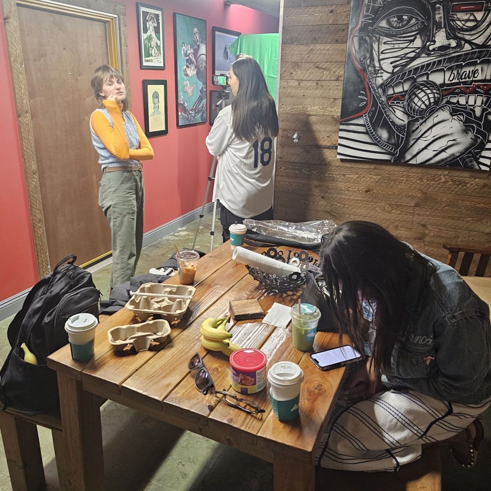Morgan Rutter talks with Emily Castro next to a VO booth while Bianca Lopez sits at a table covered in crafty.