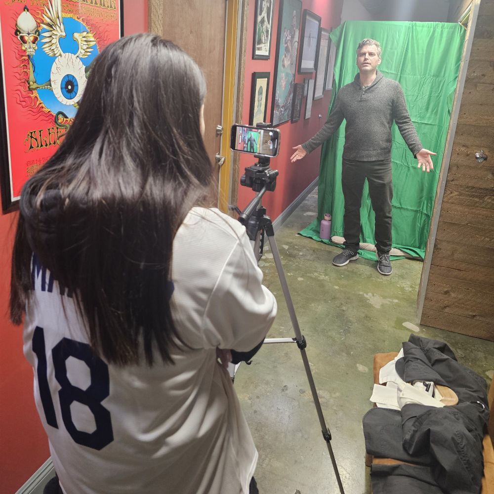 Emily Castro capturing video with a cell phone on a tripod down a long hallway. Cameron Beyl stands in front of a green screen on a door just down the hall.