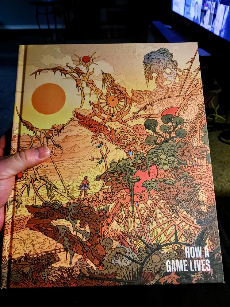 A hand holding the beautiful cover of the book, "How a Game Lives." It's huge and lovely.