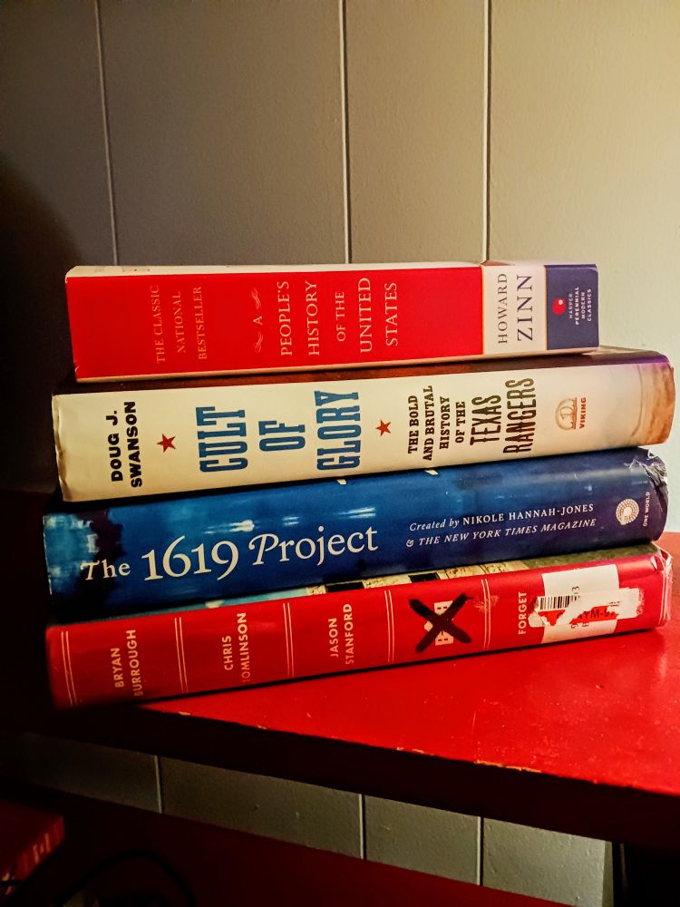 A collection of books on a table. The posing of the books are meant to convey a simple stacked feeling, almost as if they weren't stacked for a photo op. The books are historical on nature, nonfiction.
In order 
"People's History of the United States" by Howard Zinn
"Cult of Glory" by Doug J Swanson
"1619 Project" by Nikole Hannah-Jones
"Forget the Alamo" by Burrough, Tomlinson, and Stanford