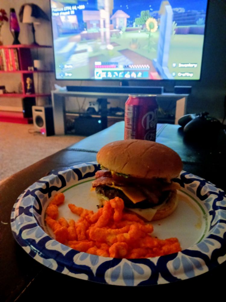 In the foreground sits a paper plate, on it is an all-American bacon cheeseburger with cheetos and a Dr. Pepper. The tv in the background is showing Minecraft, beckoning the viewer to mine for diamonds and redstone, maybe put a tiny fence around it.