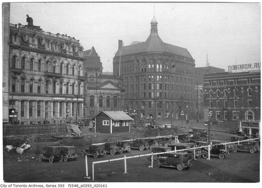 A black and white photo looking diagonally across a parking lot filled with cars from the 1920s towards an intersection. On the left is a four-storey building with mansard roofs. Next to it is the neo-classical Bank of Montreal. Diagonally across the intersection is a sandstone building that has a rounded corner with a conical roof. Photo by John Boyd Sr. Fonds 1548, Series 393, Item 20161.