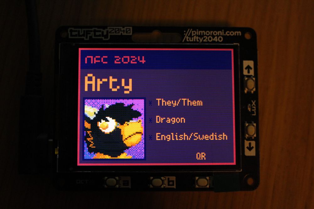 A printed circuit board with an RP2040 on the back and an LCD screen on the front. The screen displays an ID badge for NFC 2024. It displays the name Arty followed by They/Them, Dragon, English/Swedish. There is also a button to display a QR code.