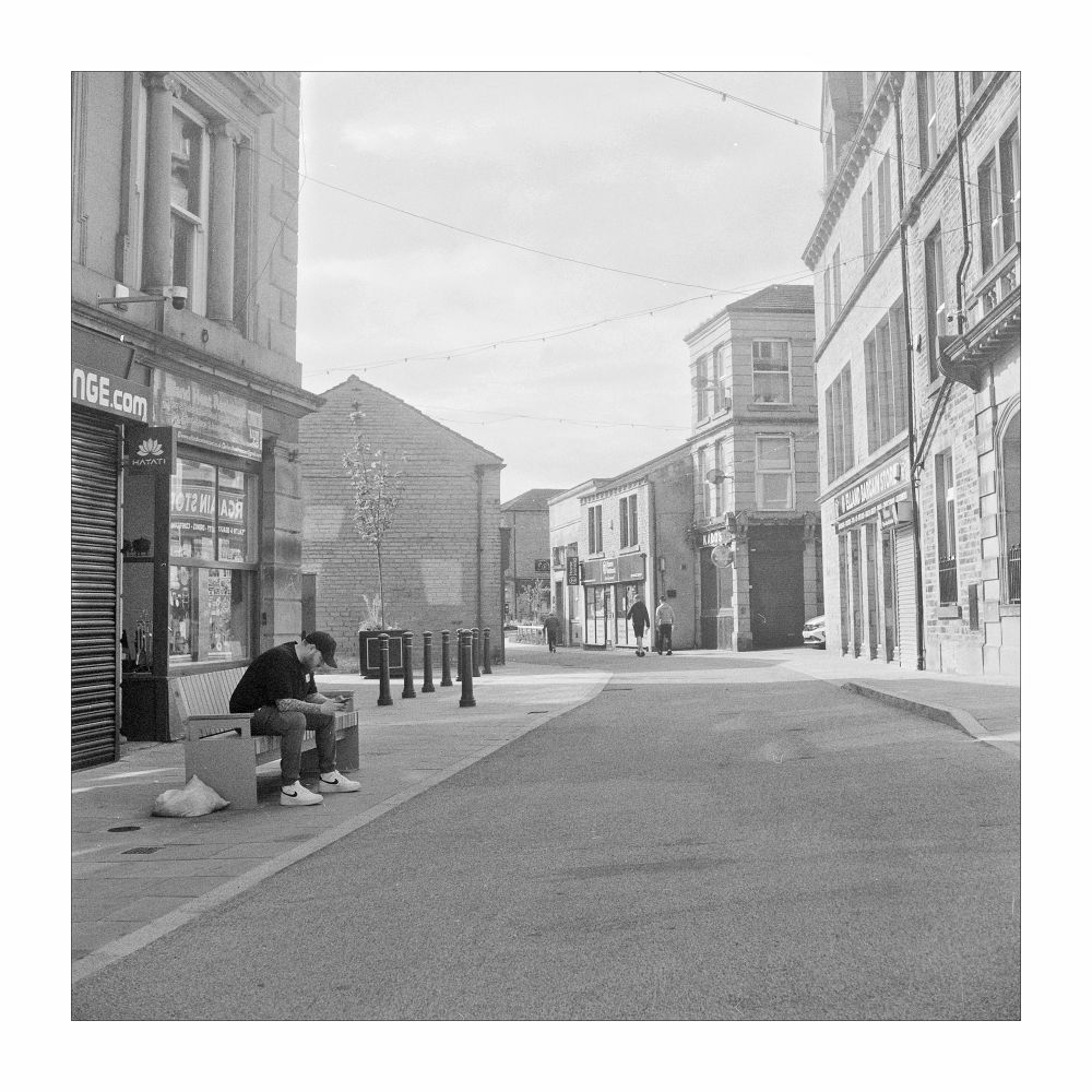 Square black and white photograph looking down a quiet high street. A man sits on a bench to the left checking his phone whilst two other gentlemen stroll through the middle of the frame 