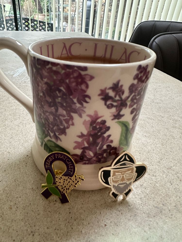 A mug of tea with a lilac blossom pattern on it, alongside lilac and Terry Pratchett pin badges.
