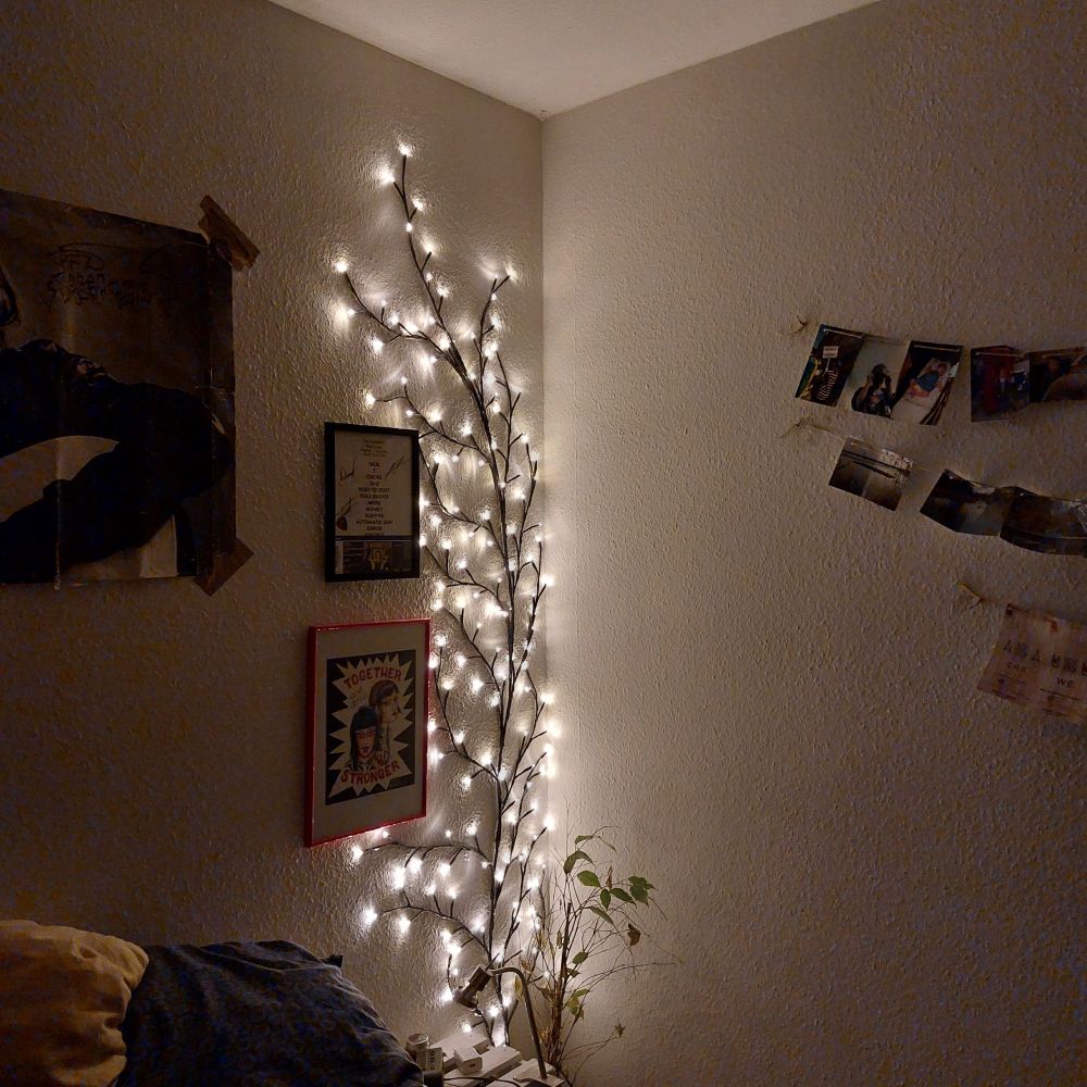 Eine Lichterkette in Baumform, die sich in einer Zimmerecke hochrankt. Davor ist ein weißer Nachttisch zu sehen, neben dem ein sterbender Ficus steht. In der rechten oberen Ecke sind einige Fotos zu sehen, in der linken die Ecke eines Kurt Cobain Posters.