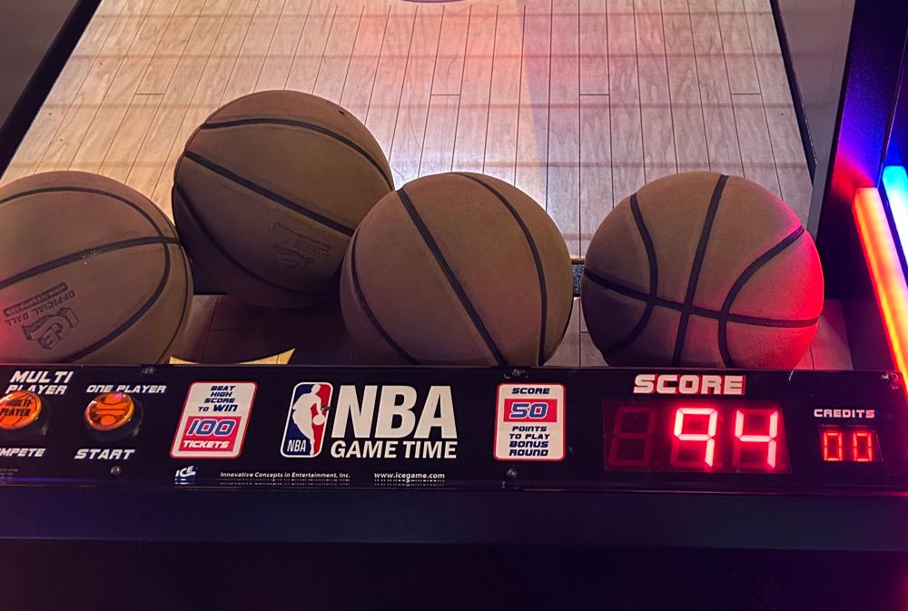 Fotografía de una máquina de arcade llamada “NBA Game Time” con un marcador de 94 puntos. Sobre la consola hay 4 balones chicos que aparentan ser balones de basquetbol.