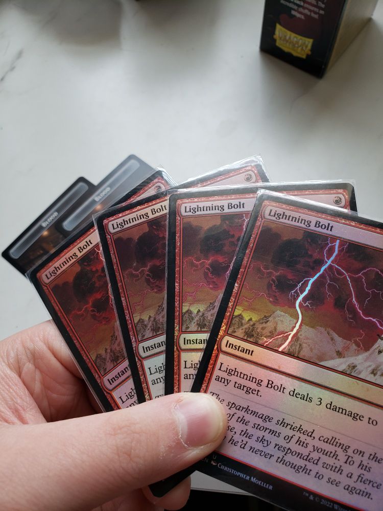 my hand gripping 4 foil copies of the Magic: the Gathering card Lightning Bolt. visible on the table behind them are two Blood tokens and a box of Dragon Shield card sleeves
