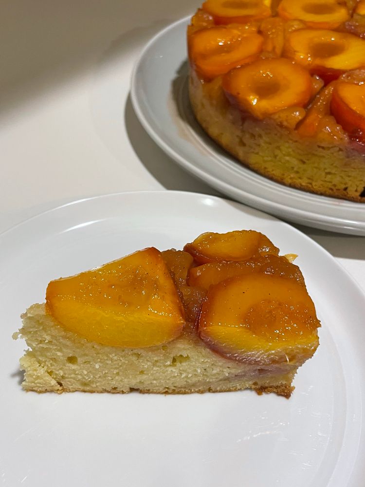 Photograph of a small piece of upside down peach cake, with glazed peaches and dripping honey, on a white dessert plate with the larger cake in the background.