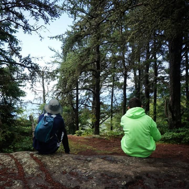 Two boys face away from the camera, sitting on rocks in the woods 