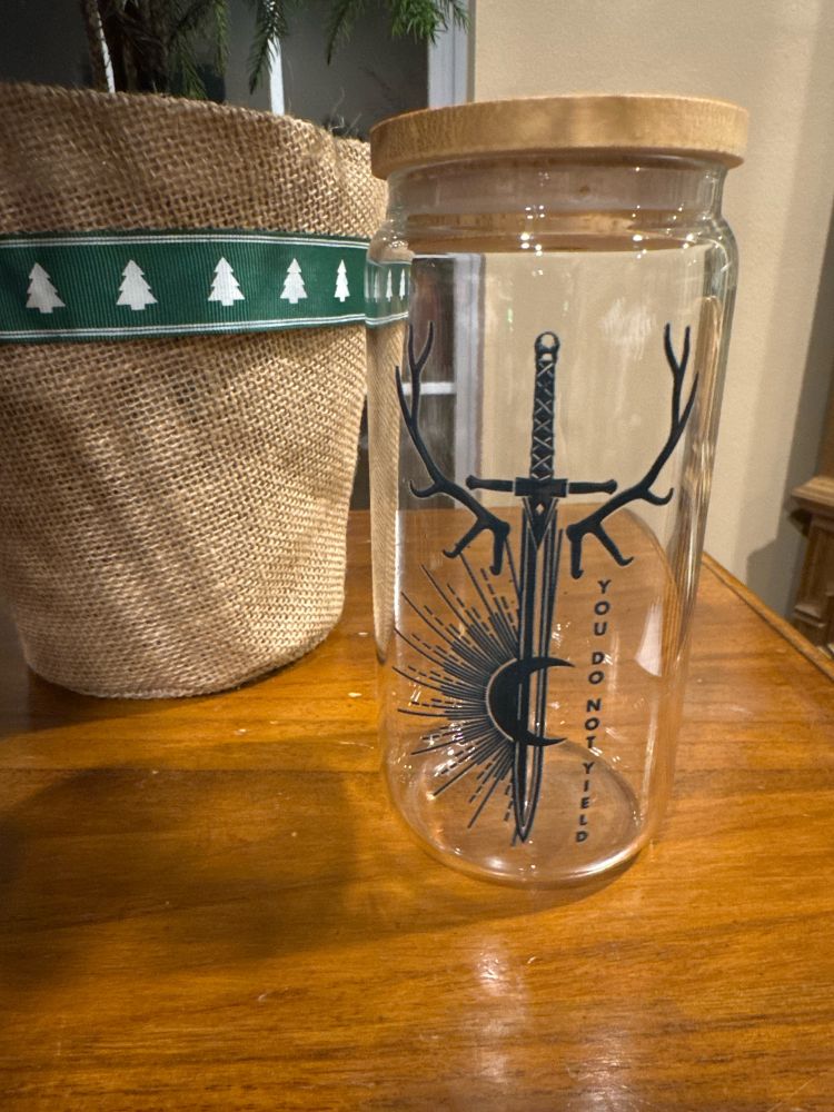 A glass jar sitting on a wooden table next to a potted plant. A sword printed on the jar pointed downward with its hilt on top. There are antlers coming out from both sides of the sword and a crescent moon over lay half sun with rays printed on the left side of the blade.
