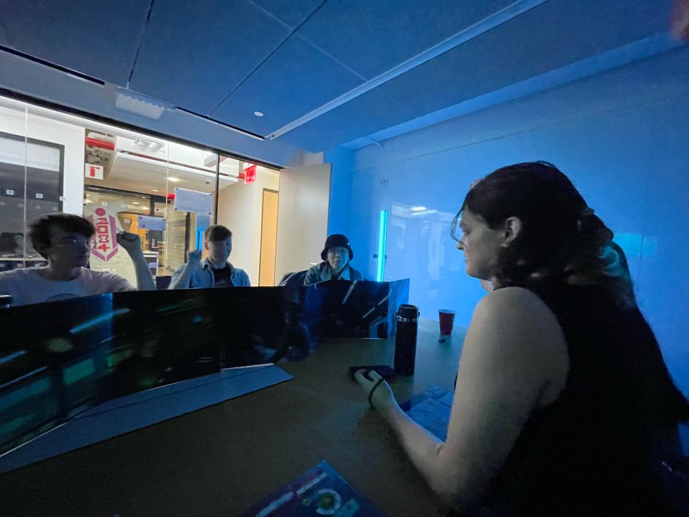 A group of game designers from the NYU Game Center Incubator playing Captain Sonar around a long table, with blue lighting and a long folding screen dividing the table.