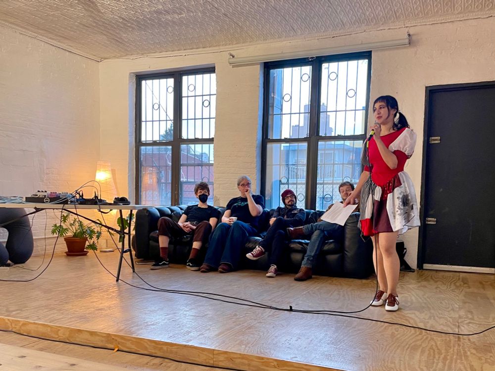 Ashley stands on a stage, speaking into a microphone as she introduces this month’s “video games reading night,” with four guest readers (mostly games journalists) sitting on a couch behind her.