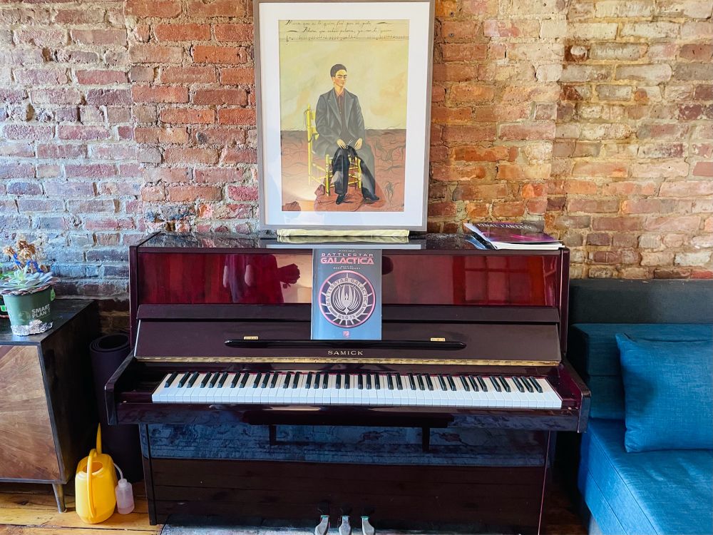 A beautiful, mahogany upright piano against an exposed brick wall. A painting of Frida Kahlo sits on top of it and a copy of the Battlestar Galactica Songbook rests on its music stand.