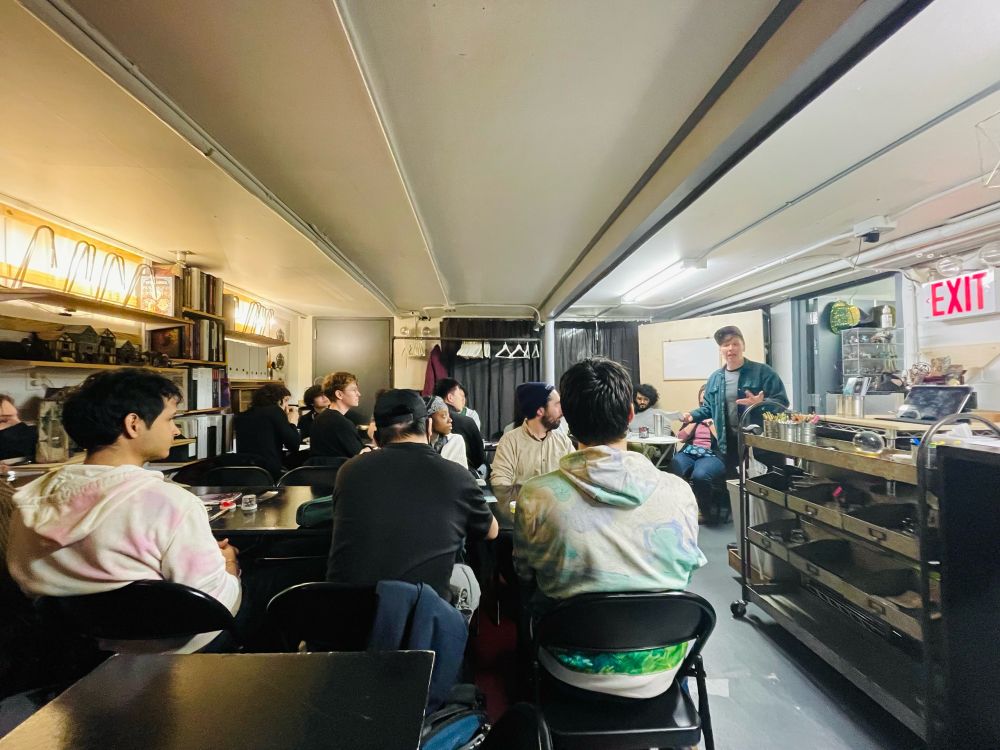 A crowded room of game designers sitting around tables in the basement of the Twenty Sided Store. They are listening to Nat Mesnard, who wears a cap and stands at the front of the room, introduce the workshop.