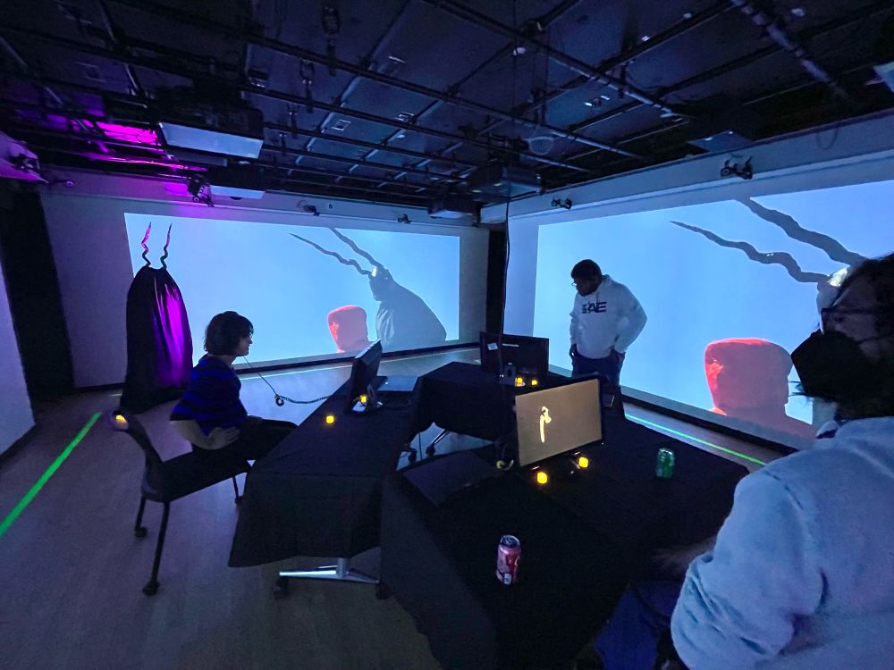 A darkened room at NYU, with video projections on several walls, a trio of computer monitors set up in the middle, and a ghostly mannequin with horns lurking in the back left 