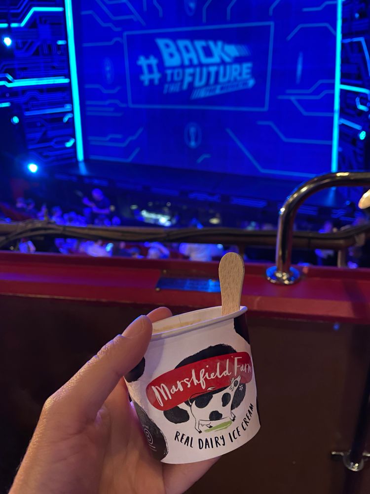 Im Theater. Im Vordergrund wird ein kleiner Eisbecher in die Kamera gehalten. Im Hintergrund sieht man die Bühne von "Back To The Future".