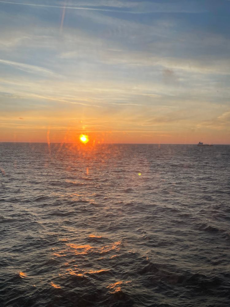 Sonnenaufgang über dem Meer (Ostsee), fotografiert durch eine Glasscheibe von einer Fähre aus. Etwas undeutlich kann man am Horizont nahe des rechten Bildrandes noch die Umrisse eines Bootes erkennen.